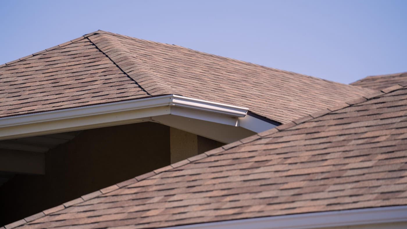 Tan asphalt shingle roof with white gutters against a clear sky